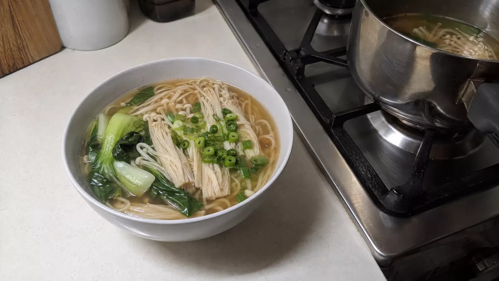 A bowl of noodle soup with bundles of enoki mushrooms, bok choy and spring onion beside a pot on a stovetop