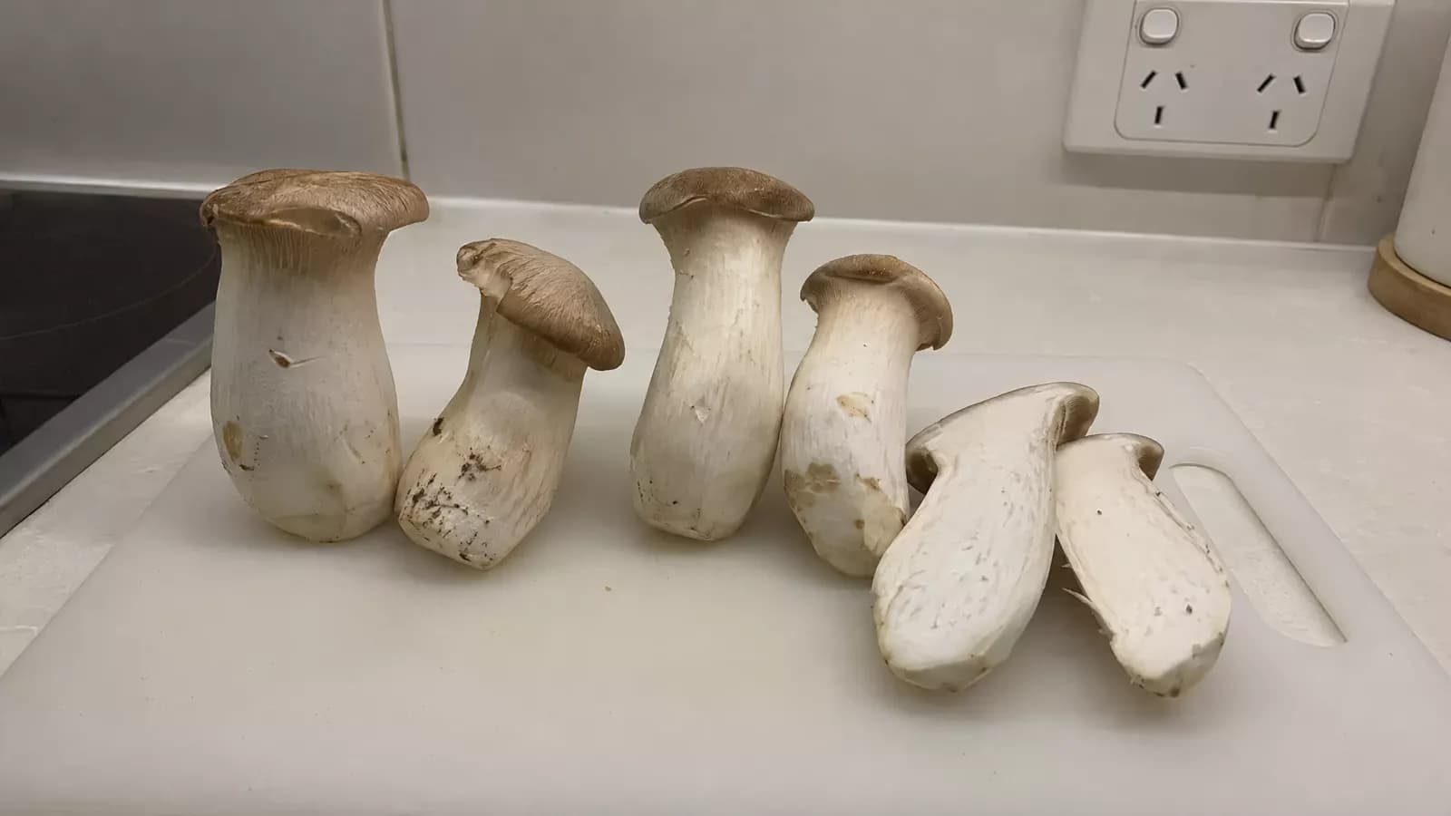 Whole king oyster mushrooms with thick white stems, and one cut in half, on a chopping board in a home kitchen